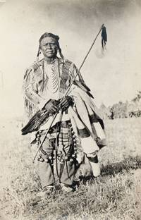 [Standing portrait of a Blackfeet man in a painted deerskin shirt, with dancing bells by his legs, holding an eagle feather fan, scalp stick and blanket]