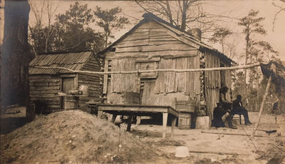 Photograph of three black men sitting outside clapboard-covered log cabin and shed