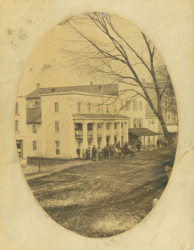Oval photograph of "Park Hotel", three storey columned clapboard structure, with group of men standing on corner outside, a waiting buckboard and other horses, others on the upper porch of the hotel